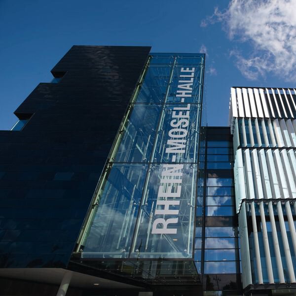 Abbildung des Veranstaltungsortes des Deutschen Nahverkehrs Tages in Karlsruhe. Die Rhein-Mosel-Halle ist von außen zu sehen mit ihrer futuristischen Glasfassade.